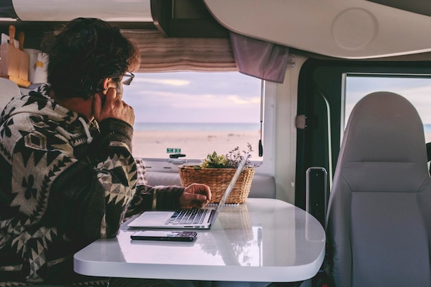 Homme travaillant à l'intérieur du camping-car assis à la table et regardant par la fenêtre la belle plage et l'océan en arrière-plan. Concept de style de vie de liberté et de travailleurs en ligne à distance