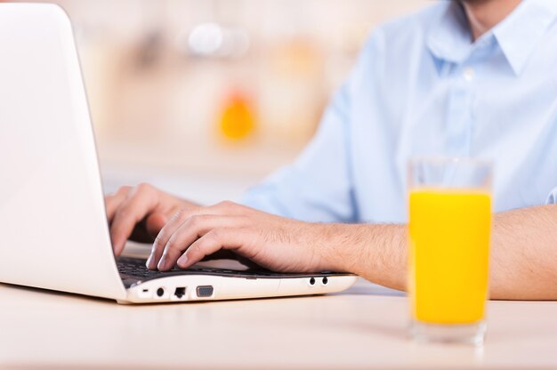 Homme travaillant sur ordinateur portable. Image recadrée d'un homme en chemise travaillant sur un ordinateur portable assis dans la cuisine avec un verre de jus d'orange au premier plan