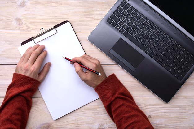 Un homme travaille sur un ordinateur portable, les mains écrivent dans un presse-papiers avec un crayon sur une table en bois, vue de dessus