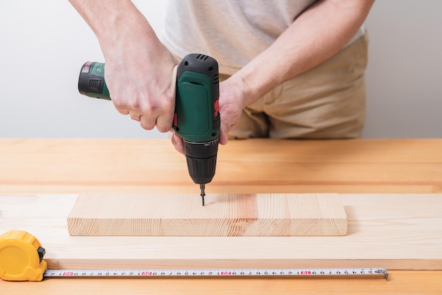 Un homme travaille avec un tournevis électrique sur une table en bois fait également des mesures avec un ruban à mesurer