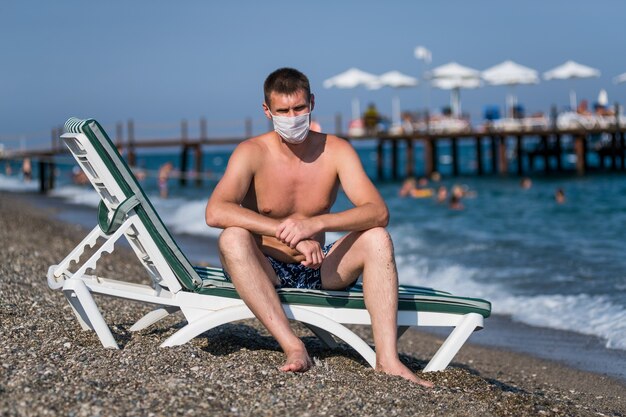 Un homme triste est assis dans une chaise longue portant un masque seul sur la plage pendant l'épidémie de grippe covid 19