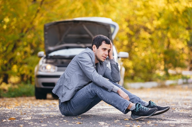 Un homme triste est assis sur la route devant une voiture cassée avec un capot ouvert