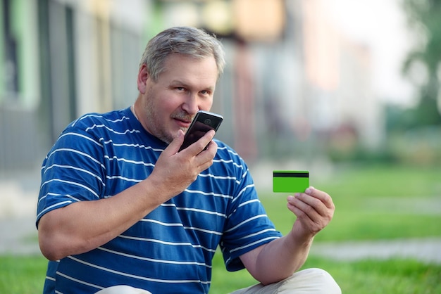 Un homme trouve des offres de commerce électronique pour acheter en ligne avec téléphone et carte de crédit sur la pelouse de la ville