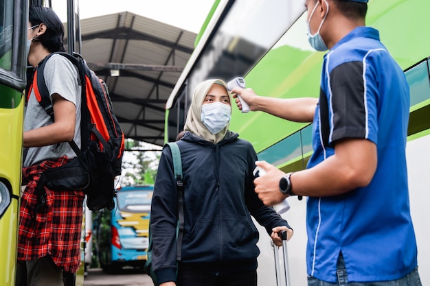 Un homme en uniforme et une casquette à l'aide d'un pistolet thermique inspecte le passager d'une femme portant un foulard et un masque avant de monter dans le bus