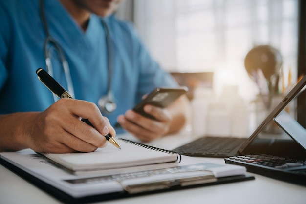 Homme en uniforme de médecin utilisant un smartphone et une tablette à technologie numérique pour périphérique de sortie et rédigeant un rapport de patient sur le bureau