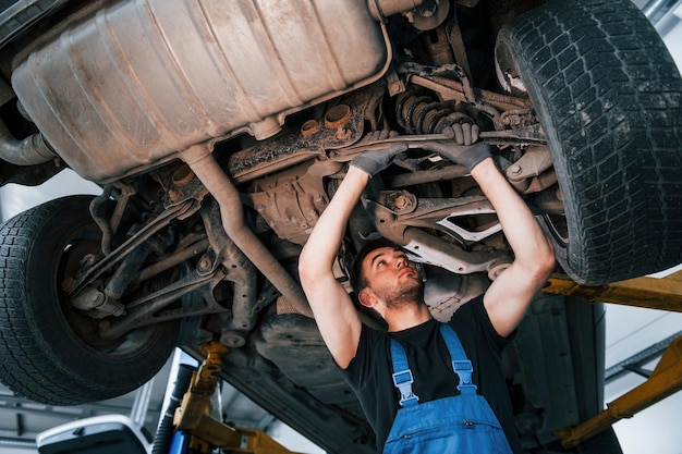 Homme en uniforme de travail debout sous la voiture et le répare à l'intérieur Conception du service automobile