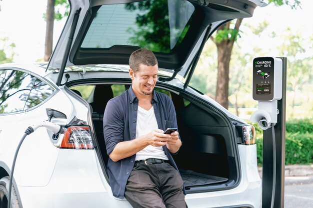 Homme utilisant une application de banque en ligne pour smartphone pour payer le chargement de la batterie d'une voiture électrique à partir d'une station de chargement de véhicules électriques pendant un voyage de vacances dans un parc national ou une forêt d'été Exalt