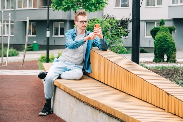 Homme utilisant communique sur un smartphone assis sur un banc sur un fond de maisons