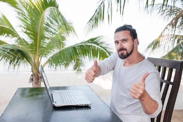 Homme Utilisant Un Ordinateur Portable Alors Qu'il était Assis Au Café De La Plage Près De Palmiers Et Montrant Les Pouces Vers Le Haut