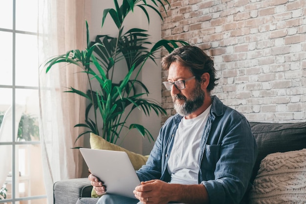 Homme utilisant un ordinateur portable assis sur un canapé dans un appartement moderne Homme de race blanche à lunettes regardant le contenu multimédia se détendre sur un canapé confortable à la maison Homme mûr travaillant à distance depuis sa maison