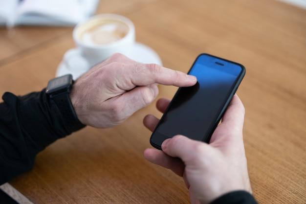 Homme utilisant son téléphone dans un café. Smartphone avec écran d'espace de copie vierge. Tasse de cappuccino ou journal plat blanc et ouvert sur table en bois. Les mains de l'homme hodling smartphone.