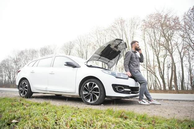 L'homme utilise un garage d'appel de téléphone portable devant le capot ouvert d'une voiture cassée sur la route dans la forêt Concept de panne de voiture