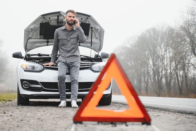 L'homme utilise un garage d'appel de téléphone portable devant le capot ouvert d'une voiture cassée sur la route dans la forêt Concept de panne de voiture