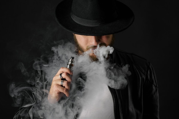 Homme avec vaping mod exhalant de la vapeur au studio noir. Gars barbu fumant une cigarette électronique pour arrêter de fumer. Concept de fumer sans vapeur et sans nicotine