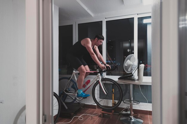 Photo homme à vélo sur l'entraîneur de machine il fait de l'exercice à la maison la nuit en jouant au jeu de course en ligne à vélo