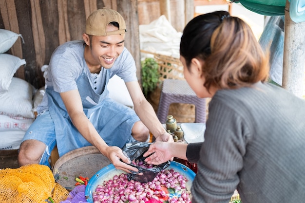 Homme vendeur tenant un sac en plastique invite les femmes acheteurs à sélectionner les échalotes au stand de légumes