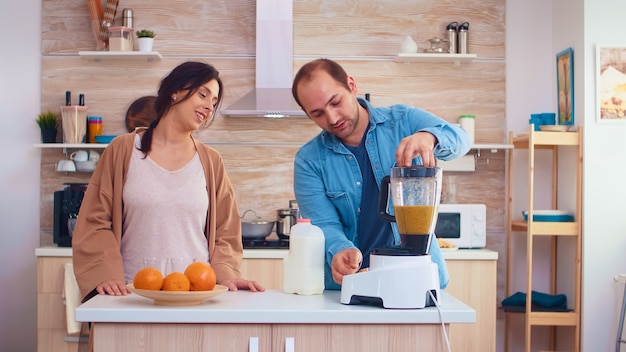 Homme versant du lait dans un mélangeur pour un smoothie frais et sain pour lui et sa femme joyeuse. Joyeuse famille faisant ensemble du jus de fruits frais et nutritif biologique sain pour le petit-déjeuner à partir de fruits frais tout en