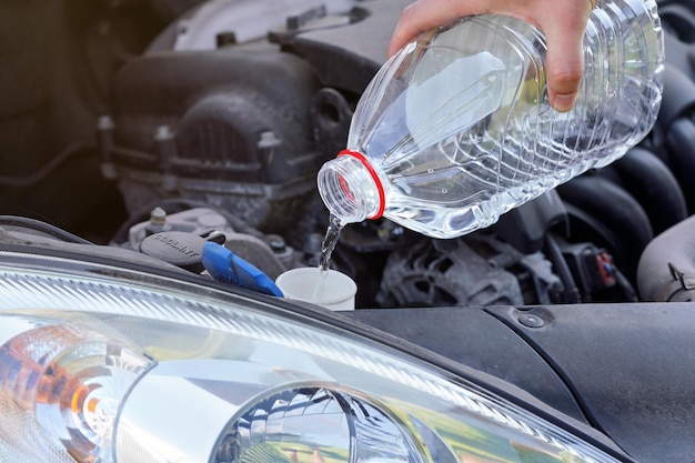 Homme versant de l'eau distillée (alternative écologique au liquide de lavage) au réservoir de lave-glace dans la voiture, détail sur la main tenant une bouteille en plastique transparent