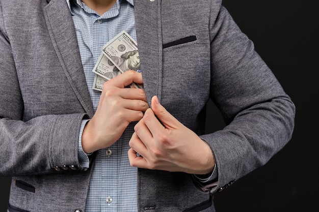 Un homme en veste met des dollars dans sa poche sur un fond noir.