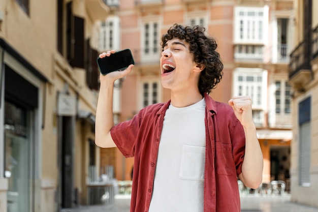 Homme en vêtements décontractés debout dans la rue à l'extérieur avec un téléphone portable à la main et se réjouit