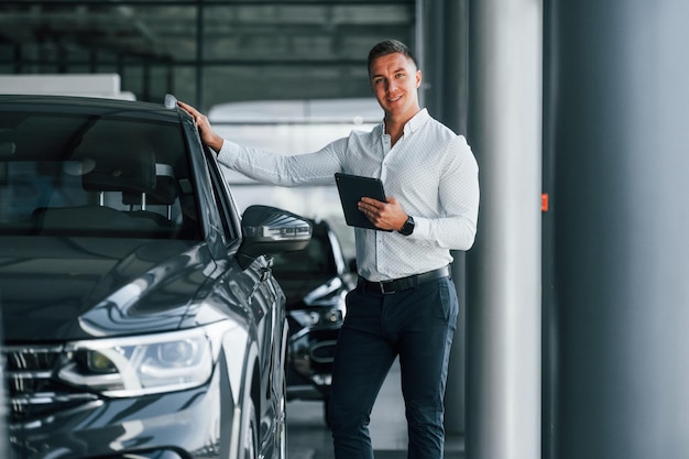 Un homme en vêtements formels se tient à l'intérieur près d'une nouvelle voiture moderne