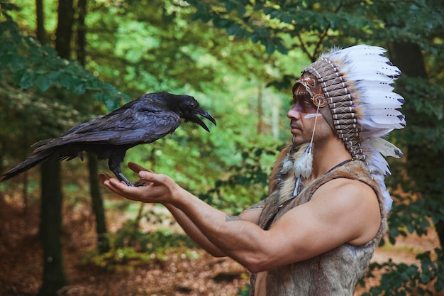 Un homme en vêtements traditionnels amérindiens tenant un corbeau dans la forêt du soir