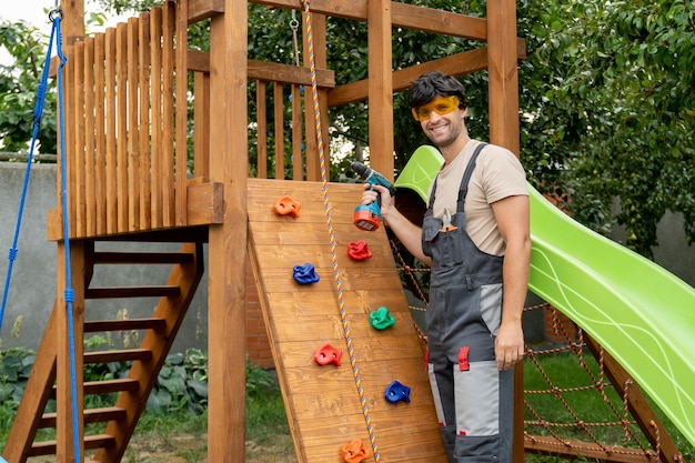 L'homme en vêtements de travail assemble un complexe de jeux en bois dans le jardin de l'arrière-cour