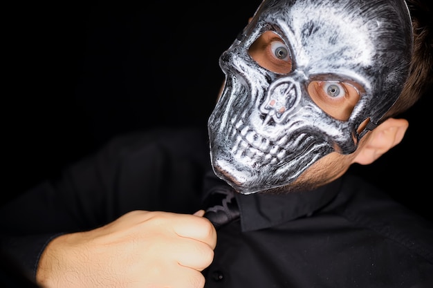 Un homme vêtu de noir se tient devant la caméra portant un masque squelette et redresse sa cravate
