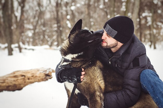 Un homme vêtu d'une veste et d'un chapeau tricoté se promène dans une forêt enneigée avec un chien Akita américain