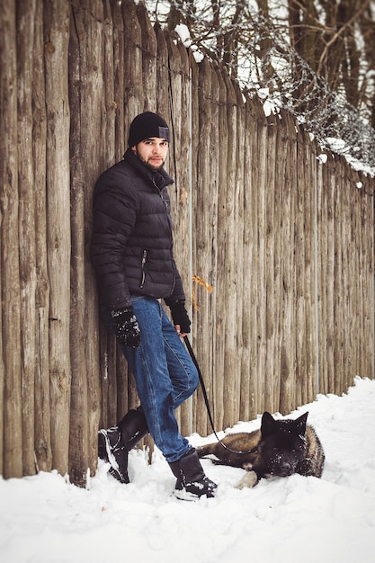 Un homme vêtu d'une veste et d'un chapeau tricoté se promène dans une forêt enneigée avec un chien Akita américain