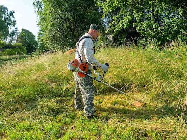 Un homme vêtu de vêtements de travail à l'aide d'une tondeuse à essence coupe l'herbe longue Zone rurale d'été entretien du territoire