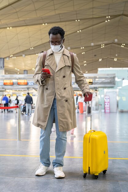 Homme voyageur afro-américain portant un masque facial pendant la pandémie de coronavirus debout au terminal de l'aéroport