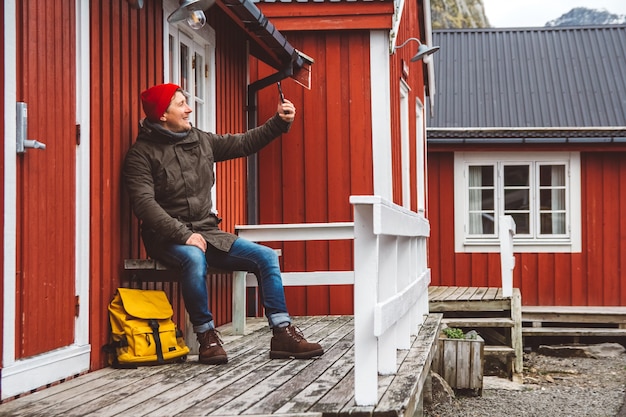 Homme de voyageur assis près de la maison en bois de couleur rouge et prenant une photo d'autoportrait