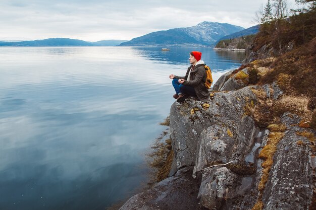 Homme de voyageur dans une position méditative assis sur un rivage rocheux sur fond de montagne et de lac