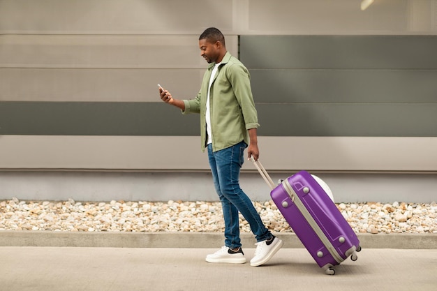 Homme voyageur noir utilisant un téléphone portable marchant avec une valise de voyage à l'extérieur