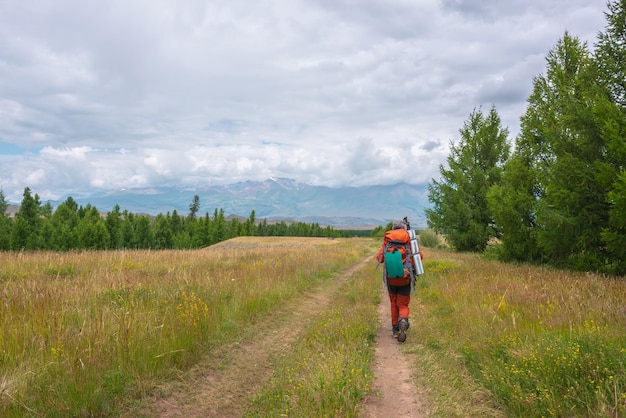 Homme voyageur en rouge avec un gros sac à dos sur le chemin de la haute chaîne de montagnes dans les nuages bas Backpacker se promène à travers la forêt jusqu'aux grandes montagnes sous un ciel nuageux Paysage dramatique avec touriste dans les montagnes