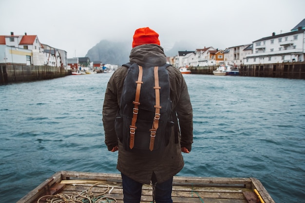 Homme voyageur avec un sac à dos portant un chapeau rouge debout sur le fond des maisons de pêcheurs, des navires, de la jetée en bois de la montagne et du lac. Concept de mode de vie de voyage. Tirez par l'arrière.