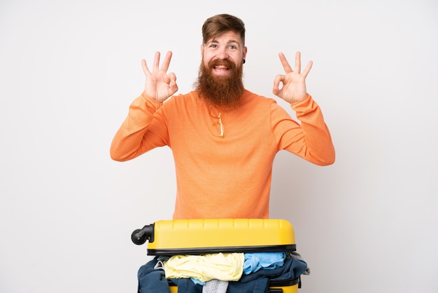 Homme voyageur avec une valise pleine de vêtements sur un mur blanc isolé montrant un signe ok avec les doigts