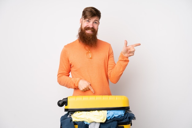 Homme voyageur avec une valise pleine de vêtements sur le mur blanc isolé pointant le doigt sur le côté