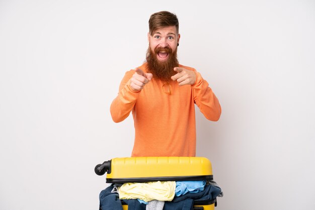 Homme voyageur avec une valise pleine de vêtements sur le mur blanc isolé pointe le doigt vers vous