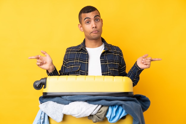 Homme voyageur avec une valise pleine de vêtements sur un mur jaune isolé pointant vers les latéraux ayant des doutes