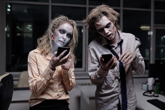 Des hommes d'affaires morts et effrayants avec de la graisse zombie sur le visage et les mains défilant dans les smartphones au bureau à minuit