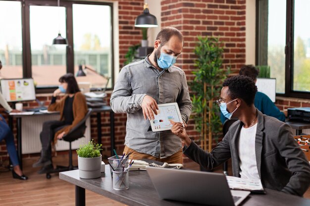 Hommes d'affaires multiraciaux comparant les statistiques marketing discutant de la stratégie de l'entreprise tout en travaillant sur un projet d'entreprise dans un bureau de démarrage de mur de briques. Équipe diversifiée avec masque médical contre covid19