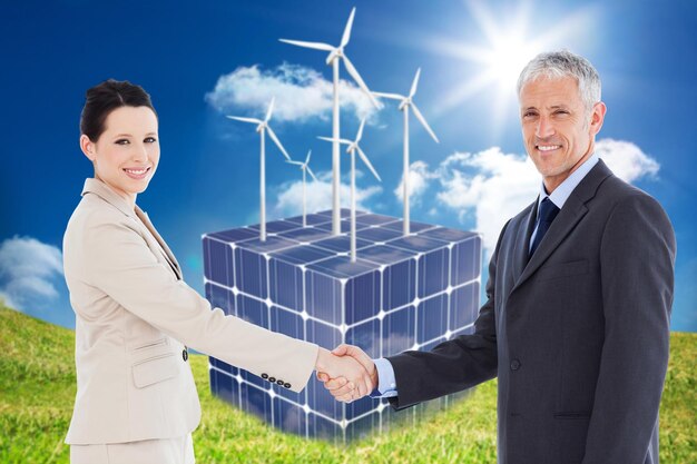 Des hommes d'affaires souriants se serrant la main tout en regardant la caméra contre des turbines sur un cube fait de panneaux solaires