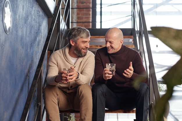 Hommes d'âge moyen positifs dans des tenues décontractées assis sur l'escalier et boire de l'alcool tout en s'amusant ensemble