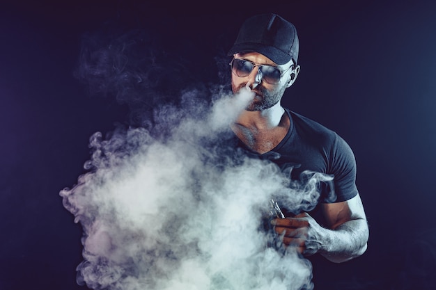 Les hommes avec la barbe dans les lunettes de soleil vaping et libère un nuage de vapeur