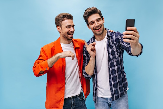 Des hommes bruns gais en jeans sourient et prennent un selfie de bonne humeur sur fond bleu Un gars en chemise à carreaux tient le téléphone