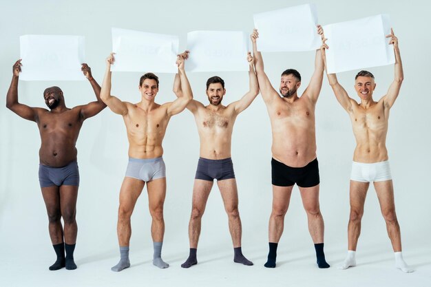 Des hommes multiethniques posant pour un ensemble de beauté positive pour le corps en édition masculine affichant des messages sur des bannières. Gars torse nu avec un âge différent et un corps portant des sous-vêtements de boxeurs