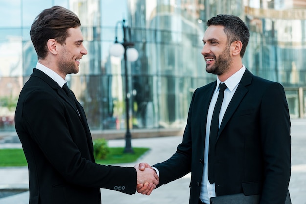 Les hommes se serrent la main. Deux jeunes hommes d'affaires confiants se serrant la main et souriant tout en se tenant à l'extérieur