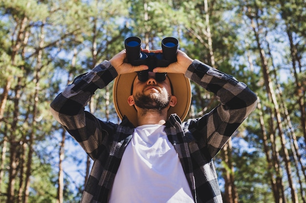 Photo les hommes touristiques dans un chapeau et une chemise à carreaux gris regarde à travers des jumelles sur un fond de forêt.